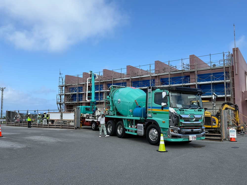A construction site in Japan featuring a large teal/green Hino concrete mixer truck (numbered 508) positioned in front of a multi-story building under renovation with scaffolding. Workers in safety gear are visible near the site entrance, and a yellow excavator is partially visible to the right. A traffic cone and a GYM-related sign are present in the foreground.