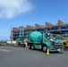 A construction site in Japan featuring a large teal/green Hino concrete mixer truck (numbered 508) positioned in front of a multi-story building under renovation with scaffolding. Workers in safety gear are visible near the site entrance, and a yellow excavator is partially visible to the right. A traffic cone and a GYM-related sign are present in the foreground.
