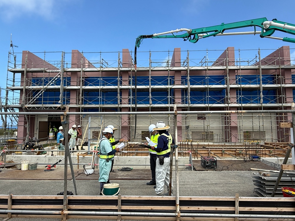 A construction site showing a multi-story building under active development, surrounded by scaffolding and safety netting. Three workers in the foreground, wearing hard hats and high-visibility vests, appear to be reviewing documents or plans. A concrete pump boom (green) is visible in the upper right, indicating active concrete pouring operations.