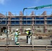 A construction site showing a multi-story building under active development, surrounded by scaffolding and safety netting. Three workers in the foreground, wearing hard hats and high-visibility vests, appear to be reviewing documents or plans. A concrete pump boom (green) is visible in the upper right, indicating active concrete pouring operations.