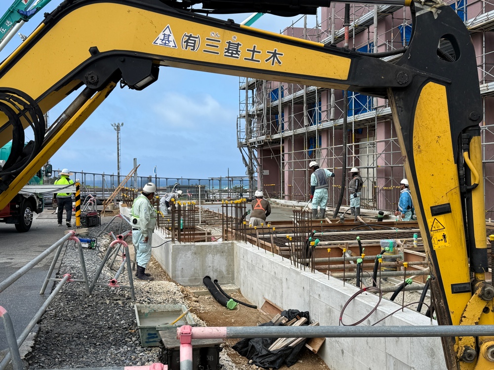 An active construction site in Japan, featuring workers performing reinforced concrete foundation work. A yellow excavator belonging to '有限会社三基土木' (Sanki Civil Engineering Co., Ltd.) is prominently visible in the foreground. Workers in white coveralls and hard hats are engaged in rebar placement and concrete formwork preparation adjacent to a multi-story building under scaffolding.