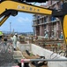 An active construction site in Japan, featuring workers performing reinforced concrete foundation work. A yellow excavator belonging to '有限会社三基土木' (Sanki Civil Engineering Co., Ltd.) is prominently visible in the foreground. Workers in white coveralls and hard hats are engaged in rebar placement and concrete formwork preparation adjacent to a multi-story building under scaffolding.