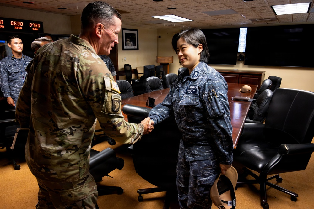 Two military personnel are shaking hands in what appears to be a command or briefing room. One individual wears a US Army multicam/OCP uniform with an airborne patch, while the other wears a blue digital camouflage uniform consistent with Asian military forces. Additional uniformed personnel are visible in the background near a large conference table.