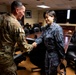 Two military personnel are shaking hands in what appears to be a command or briefing room. One individual wears a US Army multicam/OCP uniform with an airborne patch, while the other wears a blue digital camouflage uniform consistent with Asian military forces. Additional uniformed personnel are visible in the background near a large conference table.