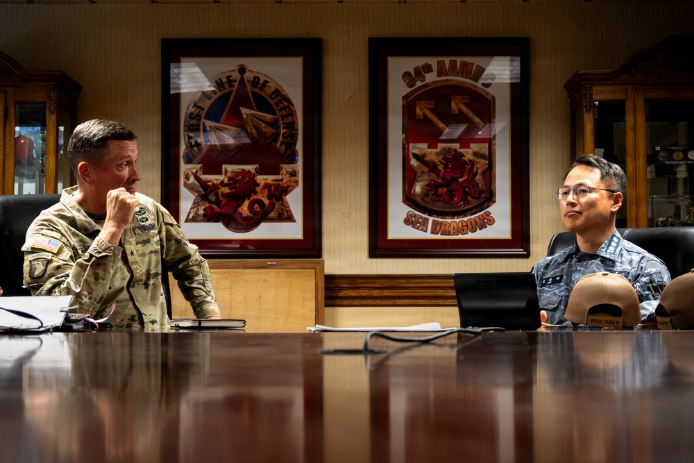 Two military personnel are seated at a conference table in what appears to be a formal military briefing room. One individual wears a U.S. Army camouflage uniform with visible rank insignia and airborne patch, while the other wears what appears to be a Republic of Korea Air Force uniform. Behind them hang two framed unit insignia plaques, one reading '94th AAMB Sea Dragons.'