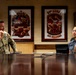 Two military personnel are seated at a conference table in what appears to be a formal military briefing room. One individual wears a U.S. Army camouflage uniform with visible rank insignia and airborne patch, while the other wears what appears to be a Republic of Korea Air Force uniform. Behind them hang two framed unit insignia plaques, one reading '94th AAMB Sea Dragons.'