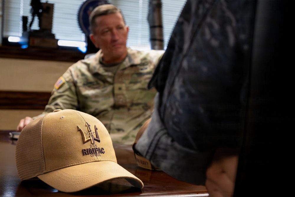 A tan baseball cap with 'RIMPAC' embroidery and a trident logo is prominently displayed in the foreground on a wooden table. In the background, a U.S. Army soldier in MultiCam (OCP) camouflage uniform with an American flag patch is seated, appearing to be in conversation. The setting appears to be a military office.
