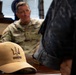 A tan baseball cap with 'RIMPAC' embroidery and a trident logo is prominently displayed in the foreground on a wooden table. In the background, a U.S. Army soldier in MultiCam (OCP) camouflage uniform with an American flag patch is seated, appearing to be in conversation. The setting appears to be a military office.