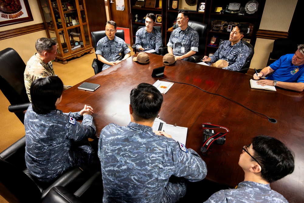 A formal military meeting is taking place around a large wooden conference table in what appears to be a senior officer's office. Multiple personnel in Republic of Korea Air Force digital camouflage uniforms are seated alongside a U.S. Army soldier in OCP uniform and a civilian in a blue polo shirt. The setting includes display cases with awards, plaques, and memorabilia in the background.