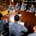 A formal military meeting is taking place around a large wooden conference table in what appears to be a senior officer's office. Multiple personnel in Republic of Korea Air Force digital camouflage uniforms are seated alongside a U.S. Army soldier in OCP uniform and a civilian in a blue polo shirt. The setting includes display cases with awards, plaques, and memorabilia in the background.