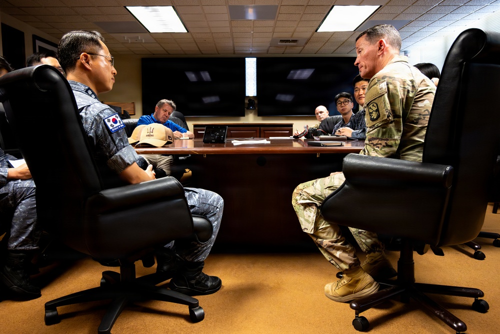 A formal military meeting is taking place in a conference room between personnel wearing Republic of Korea (ROK) Air Force uniforms and a U.S. Army soldier in MultiCam/OCP camouflage. The two primary figures are seated facing each other across a large conference table, suggesting a bilateral military discussion or briefing. Additional personnel, including civilians and uniformed individuals, are seated around the table taking notes.