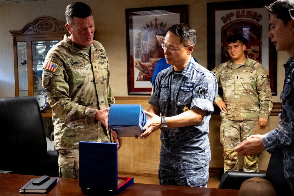 A U.S. Army officer and a Republic of Korea (ROK) military officer are exchanging a gift or ceremonial item in what appears to be a military office. The setting includes framed unit insignia on the wall, including a visible '94th AAMDC' (Army Air and Missile Defense Command) sign. Additional military personnel from both nations are present in the background observing the exchange.