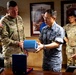 A U.S. Army officer and a Republic of Korea (ROK) military officer are exchanging a gift or ceremonial item in what appears to be a military office. The setting includes framed unit insignia on the wall, including a visible '94th AAMDC' (Army Air and Missile Defense Command) sign. Additional military personnel from both nations are present in the background observing the exchange.
