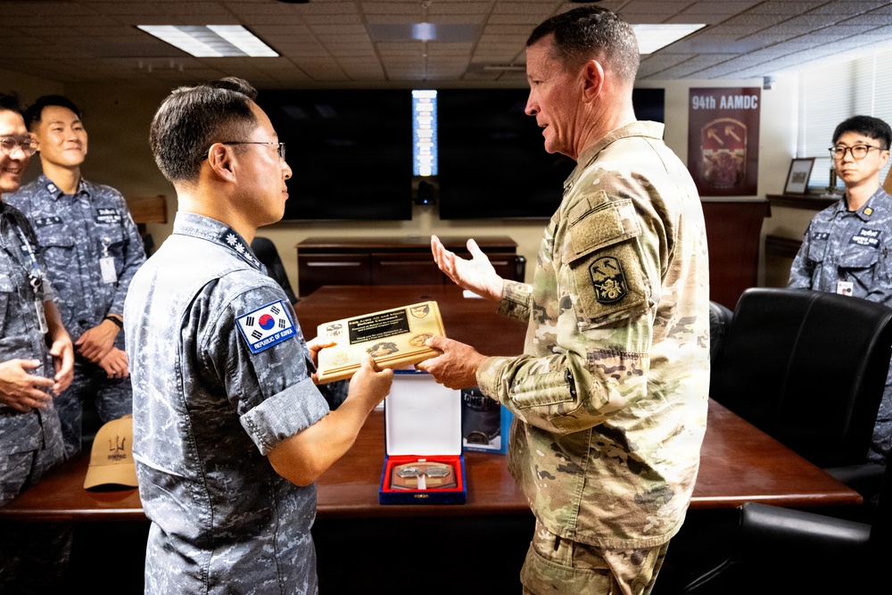 A military exchange ceremony is taking place between Republic of Korea Air Force personnel and a U.S. Army soldier, likely at a 94th Army Air and Missile Defense Command (AAMDC) facility. A Korean officer is presenting or receiving a wooden plaque/commemorative gift to/from a U.S. Army officer in MultiCam uniform. Additional Korean military personnel observe the exchange in the background.