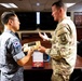 A military exchange ceremony is taking place between Republic of Korea Air Force personnel and a U.S. Army soldier, likely at a 94th Army Air and Missile Defense Command (AAMDC) facility. A Korean officer is presenting or receiving a wooden plaque/commemorative gift to/from a U.S. Army officer in MultiCam uniform. Additional Korean military personnel observe the exchange in the background.