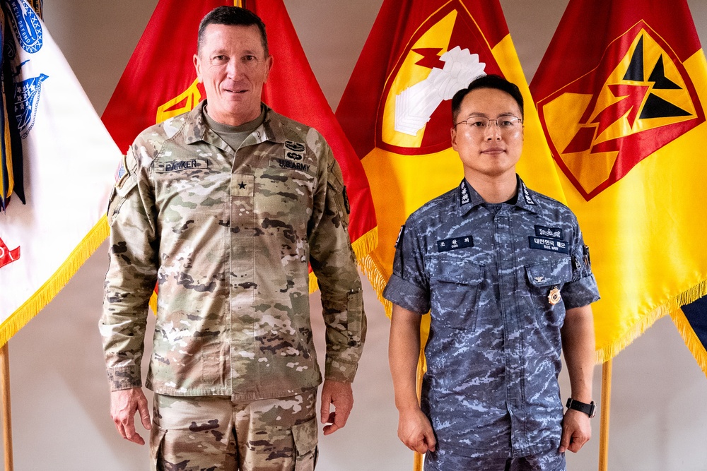 Two military personnel are posing together in front of multiple military unit flags. The individual on the left wears a U.S. Army OCP (Operational Camouflage Pattern) uniform with a name tape reading 'PARKER' and a single star indicating the rank of Brigadier General. The individual on the right wears a Republic of Korea (ROK) Air Force uniform with Korean script name tapes and the text '대한민국 공군' (Republic of Korea Air Force).