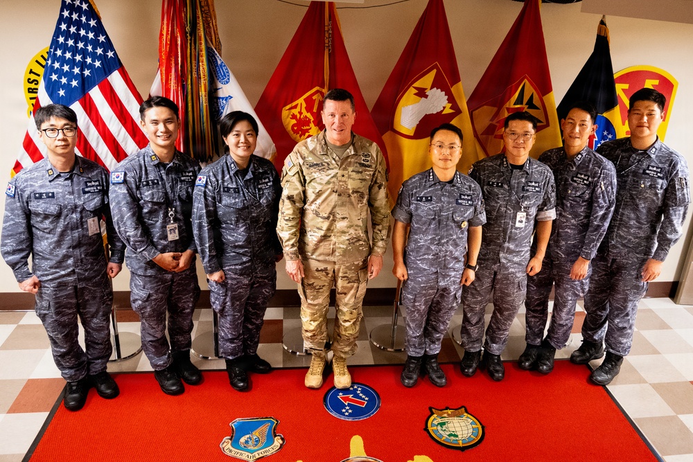 A group of eight military personnel pose for a formal photograph in front of multiple military flags including the United States flag. One individual in the center wears U.S. Army multicam (OCP) combat uniform, while the remaining seven wear what appears to be Republic of Korea Air Force digital camouflage uniforms. The setting appears to be an official military facility, with unit insignia patches visible on the floor rug.