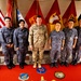 A group of eight military personnel pose for a formal photograph in front of multiple military flags including the United States flag. One individual in the center wears U.S. Army multicam (OCP) combat uniform, while the remaining seven wear what appears to be Republic of Korea Air Force digital camouflage uniforms. The setting appears to be an official military facility, with unit insignia patches visible on the floor rug.