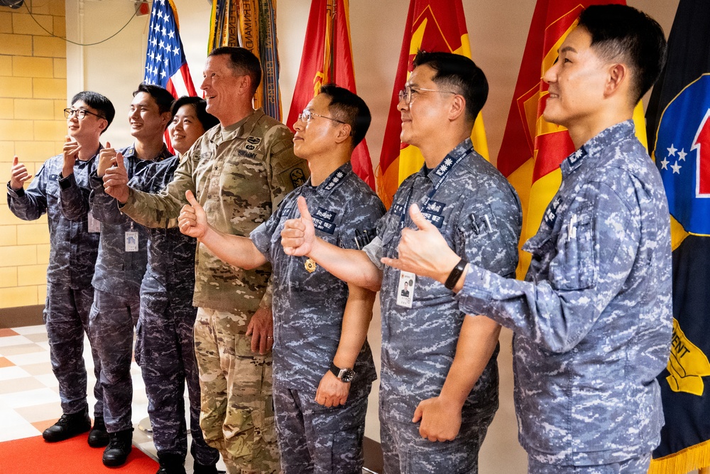 A group of military personnel, including one U.S. Army soldier in MultiCam/OCP camouflage and several individuals in blue-gray digital camouflage uniforms consistent with Republic of Korea (ROK) Air Force or Navy, are posing together giving thumbs-up gestures. They are standing in front of multiple military unit flags, including a U.S. flag and several red and yellow unit guidons. The atmosphere appears celebratory or ceremonial, suggesting a joint military meeting or visit.