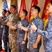 A group of military personnel, including one U.S. Army soldier in MultiCam/OCP camouflage and several individuals in blue-gray digital camouflage uniforms consistent with Republic of Korea (ROK) Air Force or Navy, are posing together giving thumbs-up gestures. They are standing in front of multiple military unit flags, including a U.S. flag and several red and yellow unit guidons. The atmosphere appears celebratory or ceremonial, suggesting a joint military meeting or visit.