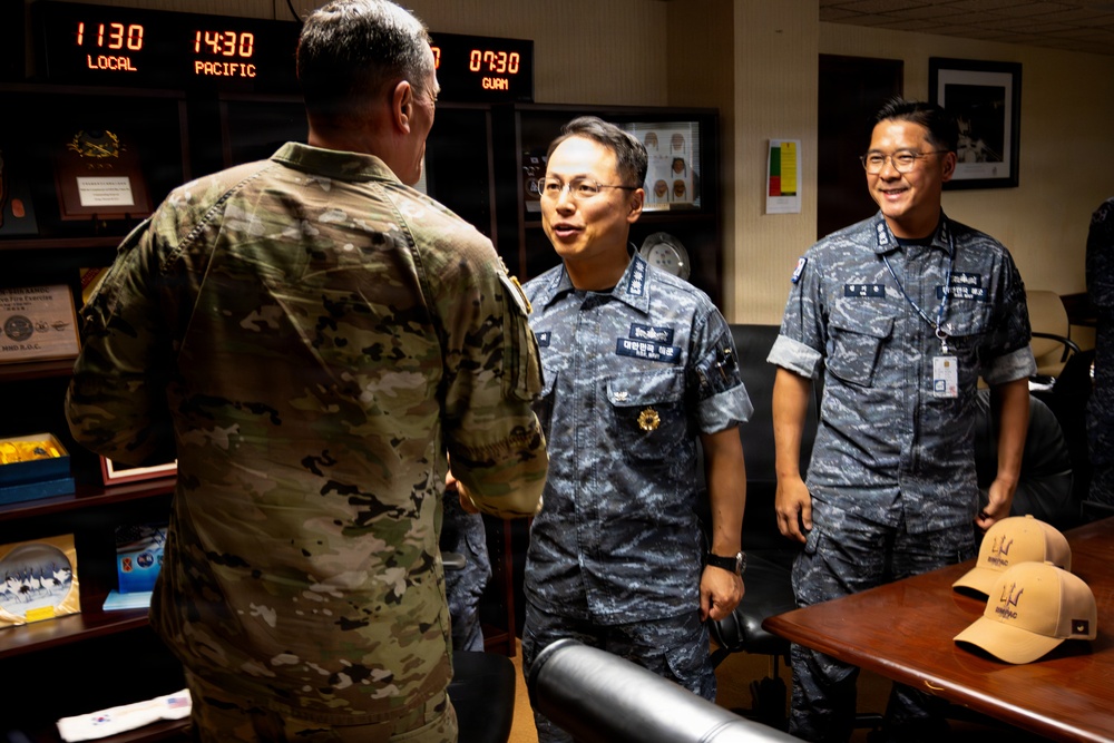 A military meeting or greeting is taking place between a U.S. Army soldier in MultiCam/OCP camouflage and two Republic of Korea (ROK) Air Force officers in their distinctive digital gray camouflage uniforms. The setting appears to be a military office or briefing room with multiple time zone clocks visible on the wall showing LOCAL (11:30), PACIFIC (14:30), and GUAM (07:30). The interaction appears cordial, with a handshake occurring between the U.S. soldier and one of the ROK officers.