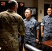 A military meeting or greeting is taking place between a U.S. Army soldier in MultiCam/OCP camouflage and two Republic of Korea (ROK) Air Force officers in their distinctive digital gray camouflage uniforms. The setting appears to be a military office or briefing room with multiple time zone clocks visible on the wall showing LOCAL (11:30), PACIFIC (14:30), and GUAM (07:30). The interaction appears cordial, with a handshake occurring between the U.S. soldier and one of the ROK officers.