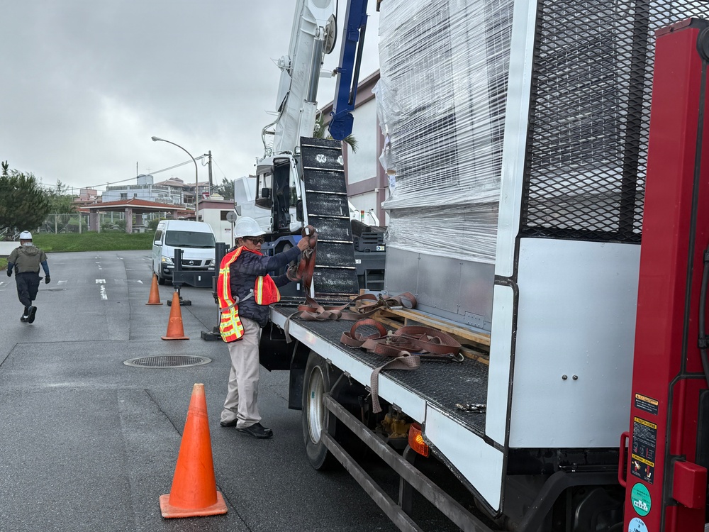 A construction or delivery worker wearing a high-visibility orange safety vest and white hard hat is working on a flatbed truck, handling cargo straps near a large plastic-wrapped load. The scene takes place on a wet road surface, suggesting recent rainfall, with traffic cones placed around the vehicle for safety. A second worker in a hard hat is visible walking in the background near a white van.