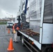 A construction or delivery worker wearing a high-visibility orange safety vest and white hard hat is working on a flatbed truck, handling cargo straps near a large plastic-wrapped load. The scene takes place on a wet road surface, suggesting recent rainfall, with traffic cones placed around the vehicle for safety. A second worker in a hard hat is visible walking in the background near a white van.