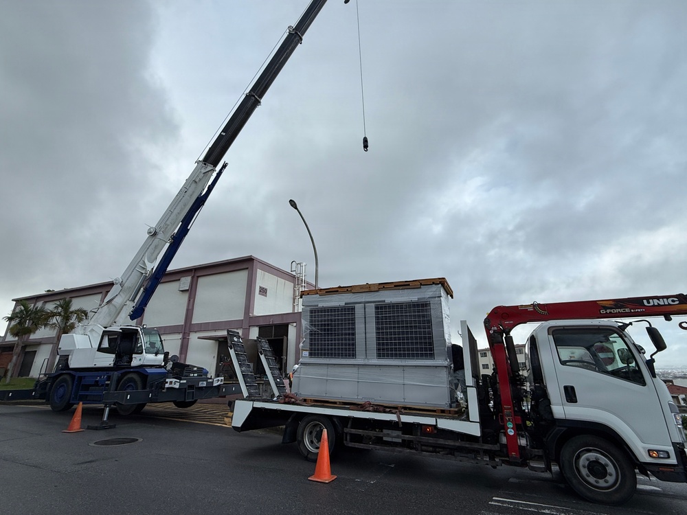 A large HVAC unit or industrial equipment is being prepared for crane lifting from a flatbed truck in a commercial or institutional parking area. A mobile crane with an extended boom is positioned alongside a white flatbed truck branded 'UNIC' with an attached knuckle boom crane. The operation is taking place under overcast skies near a pink-and-white commercial building.