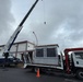 A large HVAC unit or industrial equipment is being prepared for crane lifting from a flatbed truck in a commercial or institutional parking area. A mobile crane with an extended boom is positioned alongside a white flatbed truck branded 'UNIC' with an attached knuckle boom crane. The operation is taking place under overcast skies near a pink-and-white commercial building.