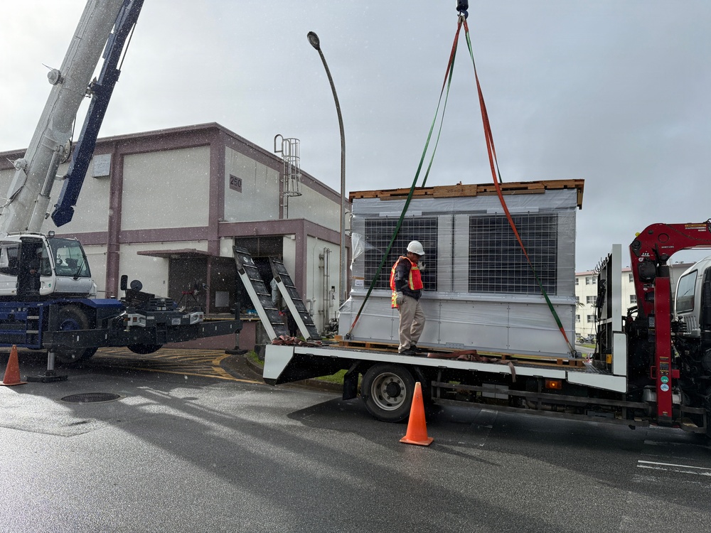 A large HVAC or rooftop air conditioning unit is being lifted by a crane from a flatbed truck in a wet, overcast outdoor setting. A worker in a hard hat and high-visibility vest stands on the truck bed, supervising or guiding the lift operation. The scene appears to take place in an industrial or institutional facility area, with a building numbered '250' visible in the background.