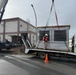 A large HVAC or rooftop air conditioning unit is being lifted by a crane from a flatbed truck in a wet, overcast outdoor setting. A worker in a hard hat and high-visibility vest stands on the truck bed, supervising or guiding the lift operation. The scene appears to take place in an industrial or institutional facility area, with a building numbered '250' visible in the background.