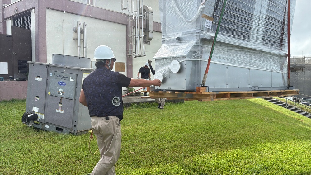 Two workers in safety gear are handling large HVAC equipment on a grassy area adjacent to a commercial or institutional building. A large unit, still wrapped in protective plastic sheeting, appears to be a newly delivered chiller or air handling unit staged on wooden pallets. The smaller unit to the left is a Carrier AquaSnap packaged HVAC unit.