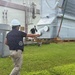 Two workers in safety gear are handling large HVAC equipment on a grassy area adjacent to a commercial or institutional building. A large unit, still wrapped in protective plastic sheeting, appears to be a newly delivered chiller or air handling unit staged on wooden pallets. The smaller unit to the left is a Carrier AquaSnap packaged HVAC unit.