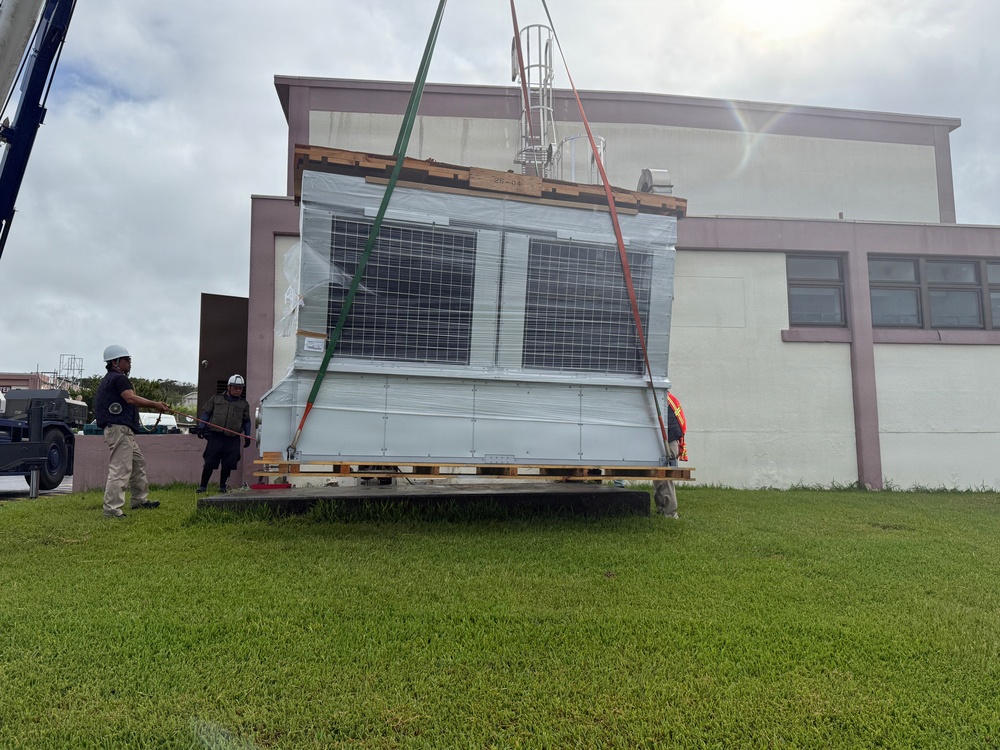 A large piece of equipment, appearing to be a solar panel array or HVAC unit, is being lifted by a crane using green and red rigging straps. Two workers wearing hard hats are guiding the load near a two-story building with a grassy foreground. The equipment is wrapped in protective plastic sheeting and mounted on a wooden pallet.