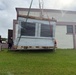 A large piece of equipment, appearing to be a solar panel array or HVAC unit, is being lifted by a crane using green and red rigging straps. Two workers wearing hard hats are guiding the load near a two-story building with a grassy foreground. The equipment is wrapped in protective plastic sheeting and mounted on a wooden pallet.