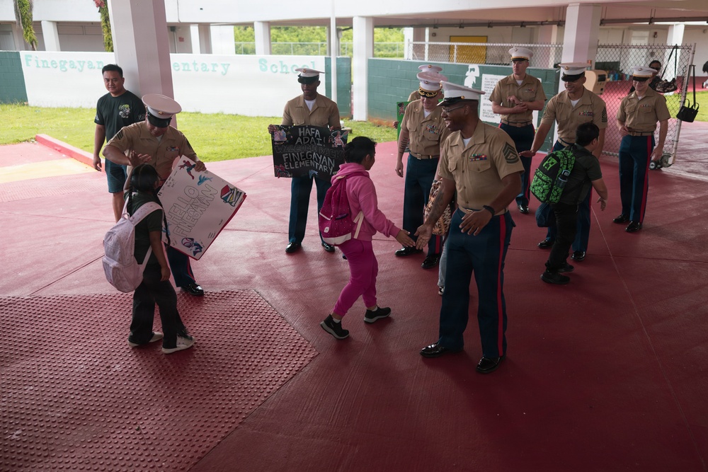 U.S. Marines in service uniforms are greeting elementary school children at Finegayan Elementary School, likely on Guam based on the 'Hafa Adai' sign visible in the scene. The Marines are forming a welcoming line and interacting warmly with students arriving at school. One Marine is holding a handmade welcome sign while another is giving a high-five to a child in a pink outfit.