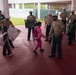 U.S. Marines in service uniforms are greeting elementary school children at Finegayan Elementary School, likely on Guam based on the 'Hafa Adai' sign visible in the scene. The Marines are forming a welcoming line and interacting warmly with students arriving at school. One Marine is holding a handmade welcome sign while another is giving a high-five to a child in a pink outfit.