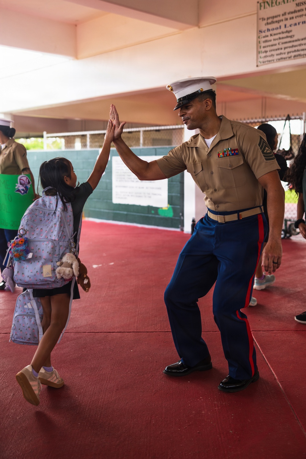 A U.S. Marine in dress uniform gives a high-five to a young girl carrying a backpack at what appears to be a school setting. The Marine is wearing the characteristic khaki shirt, blue trousers with red stripe, white cover (hat), and multiple service ribbons. The interaction appears to be part of a community outreach or school visit event.