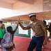 A U.S. Marine in dress uniform gives a high-five to a young girl carrying a backpack at what appears to be a school setting. The Marine is wearing the characteristic khaki shirt, blue trousers with red stripe, white cover (hat), and multiple service ribbons. The interaction appears to be part of a community outreach or school visit event.