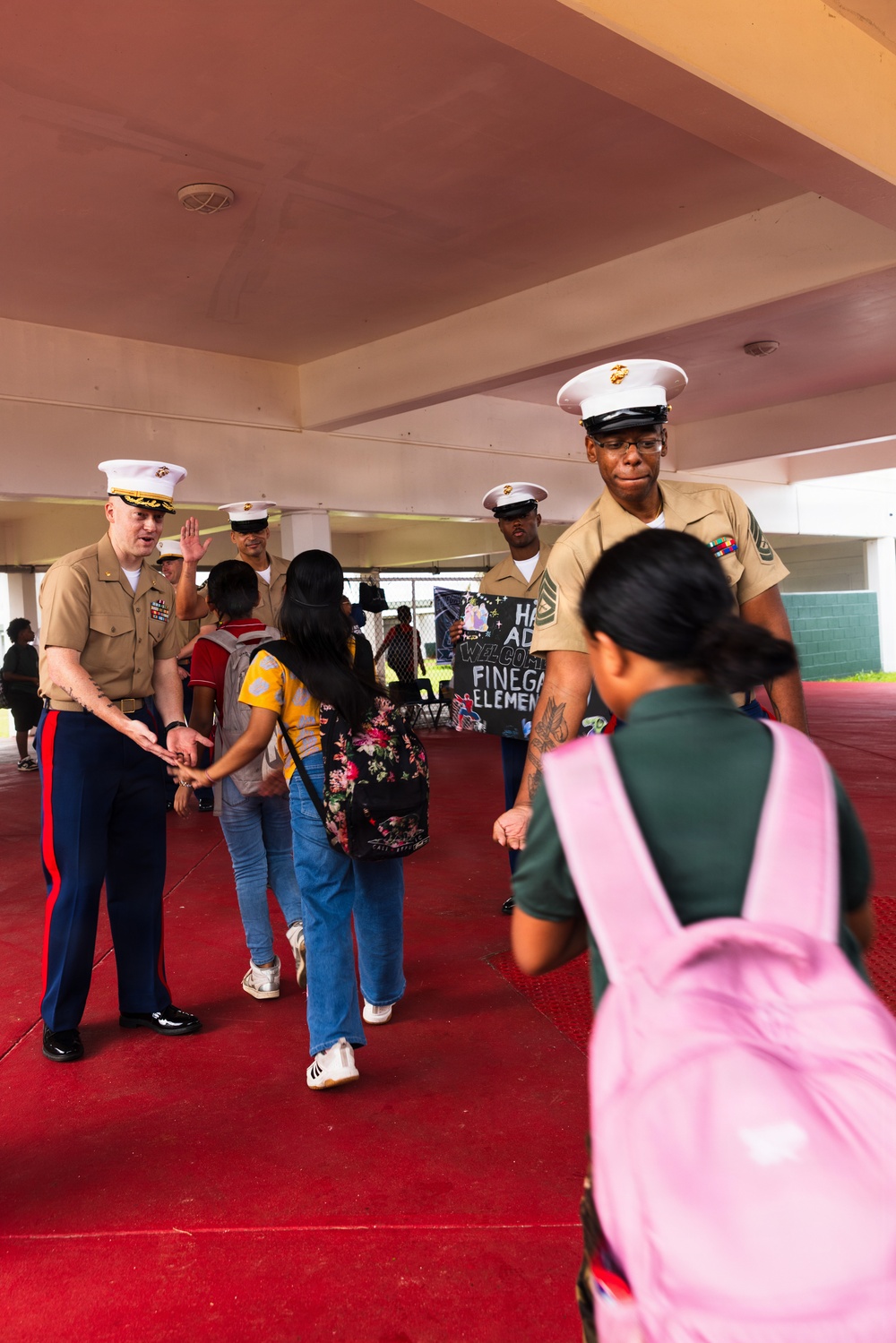 U.S. Marines in service uniforms (tan shirts, blue trousers with red stripe, white covers) are greeting children at what appears to be a school welcome event. A red carpet is laid out at the entrance, and a handwritten sign in the background references 'Finegan Elementary.' The Marines are shaking hands and interacting warmly with students arriving with backpacks.