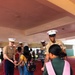 U.S. Marines in service uniforms (tan shirts, blue trousers with red stripe, white covers) are greeting children at what appears to be a school welcome event. A red carpet is laid out at the entrance, and a handwritten sign in the background references 'Finegan Elementary.' The Marines are shaking hands and interacting warmly with students arriving with backpacks.