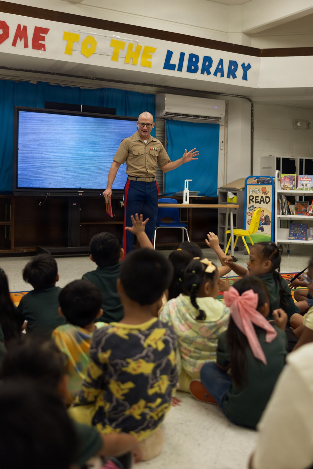 A U.S. Marine in service uniform is presenting or reading to a group of young children seated on the floor in a school library. Several children have their hands raised, indicating active engagement with the presenter. The setting appears to be a community outreach or literacy event.