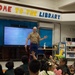 A U.S. Marine in service uniform is presenting or reading to a group of young children seated on the floor in a school library. Several children have their hands raised, indicating active engagement with the presenter. The setting appears to be a community outreach or literacy event.