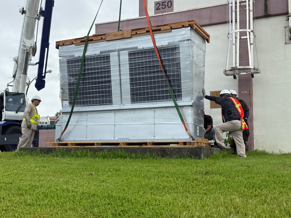 Workers in safety gear are installing or positioning a large HVAC or air conditioning unit next to a building marked with the number '250'. A crane on the left is being used to lift and maneuver the equipment, which is still partially wrapped in protective plastic sheeting and secured to wooden pallets. Three workers wearing hard hats and high-visibility vests are guiding the unit into place.