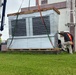 Workers in safety gear are installing or positioning a large HVAC or air conditioning unit next to a building marked with the number '250'. A crane on the left is being used to lift and maneuver the equipment, which is still partially wrapped in protective plastic sheeting and secured to wooden pallets. Three workers wearing hard hats and high-visibility vests are guiding the unit into place.