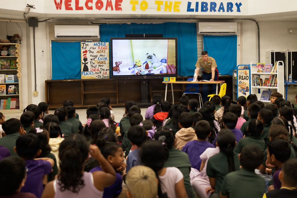 A person in military uniform is presenting a story to a large group of elementary school children in a school library. The presenter is leaning over a table near a large display screen showing an illustrated children's book. The room is decorated with colorful educational signage, including a Dr. Seuss quote and a 'Welcome to the Library' banner.