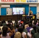 A person in military uniform is presenting a story to a large group of elementary school children in a school library. The presenter is leaning over a table near a large display screen showing an illustrated children's book. The room is decorated with colorful educational signage, including a Dr. Seuss quote and a 'Welcome to the Library' banner.