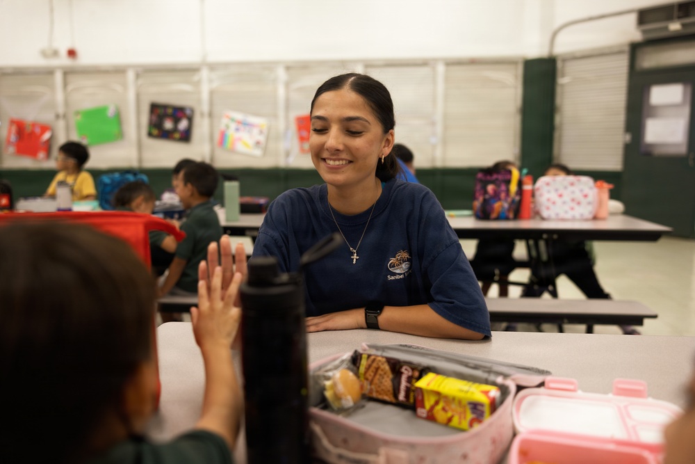 A young woman wearing a navy blue 'Sanibel' t-shirt and a cross necklace sits at a cafeteria table, smiling warmly at a child who is raising their hand toward her in what appears to be a high-five gesture. The setting appears to be a school cafeteria or multipurpose room with several children visible in the background. The scene conveys a positive, mentoring or supervisory interaction during what appears to be a lunch or snack period.