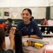 A young woman wearing a navy blue 'Sanibel' t-shirt and a cross necklace sits at a cafeteria table, smiling warmly at a child who is raising their hand toward her in what appears to be a high-five gesture. The setting appears to be a school cafeteria or multipurpose room with several children visible in the background. The scene conveys a positive, mentoring or supervisory interaction during what appears to be a lunch or snack period.